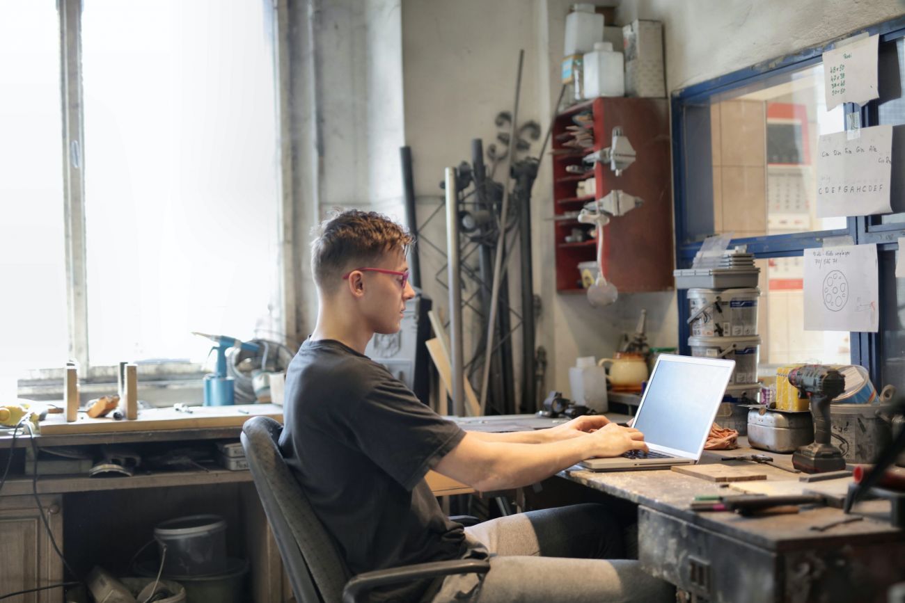 Junger Mann in einer Werkstatt, an einer Werkbank schreibt auf einem Laptop