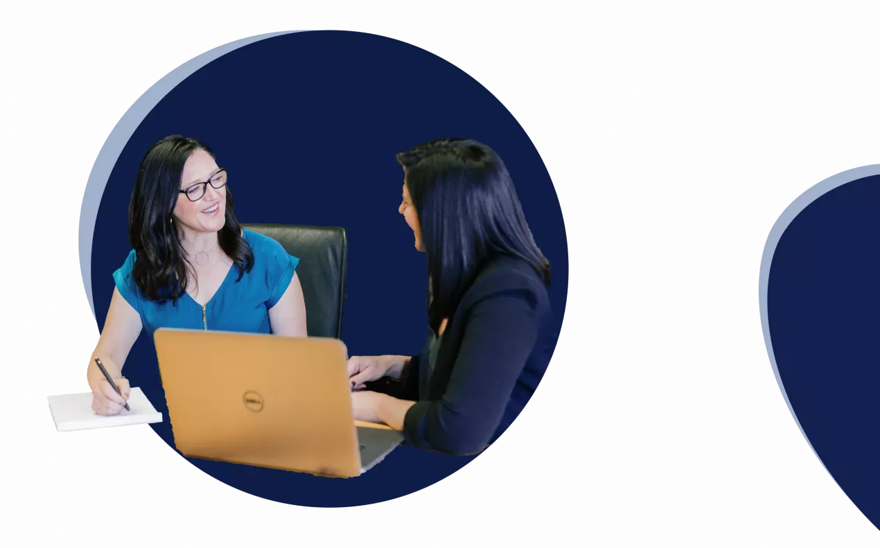 Zwei Frauen am Tisch unterhalten sich, auf dem Tisch ein Laptop und ein Notizblock.