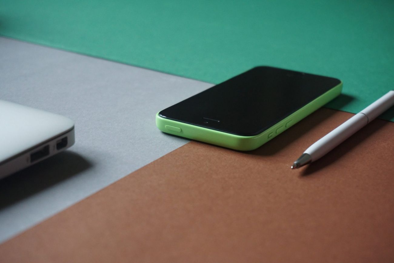 Grün, Braune Fläche, darauf ein Handy, ein Stift und die Ecke eines Laptop.
